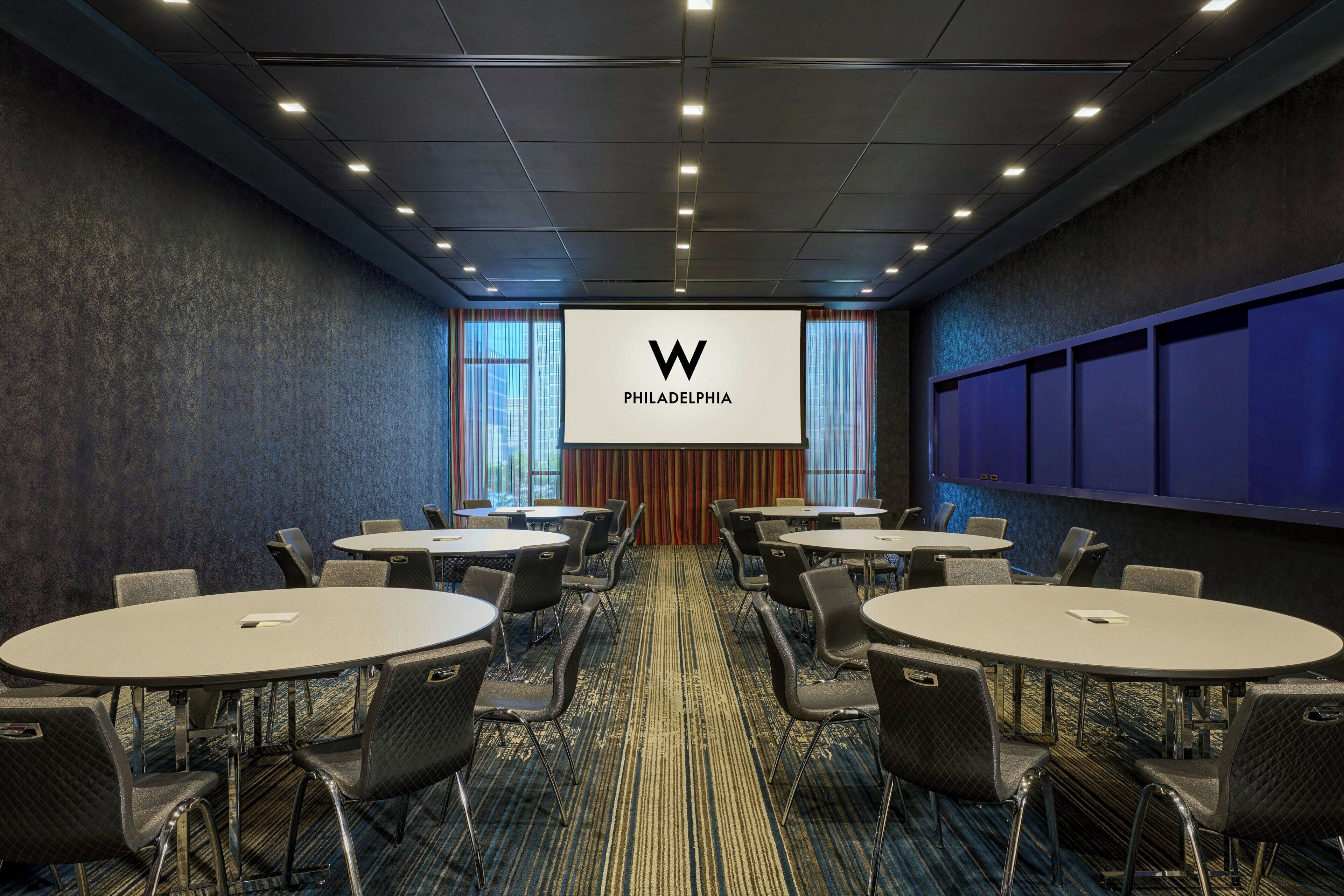 A conference room with multiple tables and chairs, a large screen displaying a logo, and a carpeted floor.