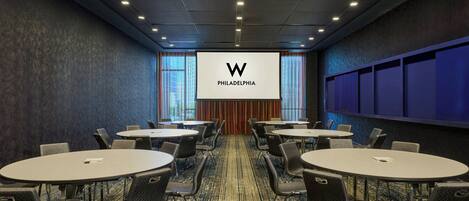 A conference room with multiple tables and chairs, a large screen displaying a logo, and a carpeted floor.