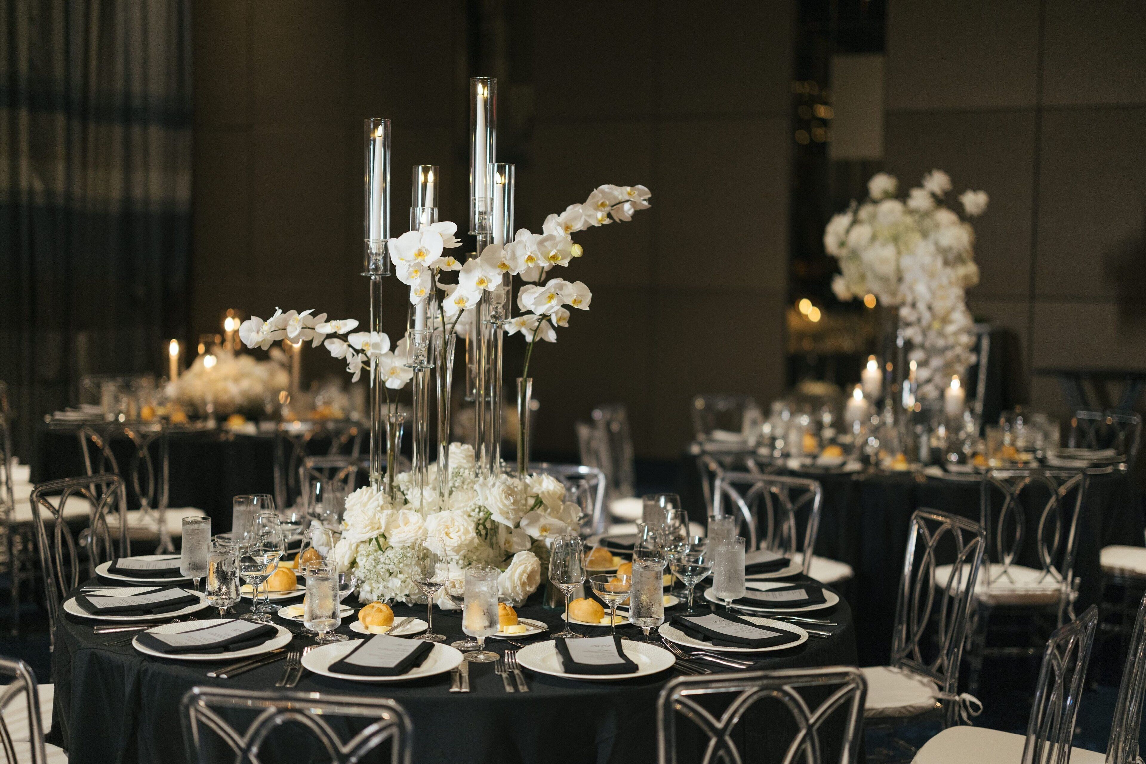 A formal dining setup with tables set for multiple guests, featuring clear glassware, white flowers, and candles.