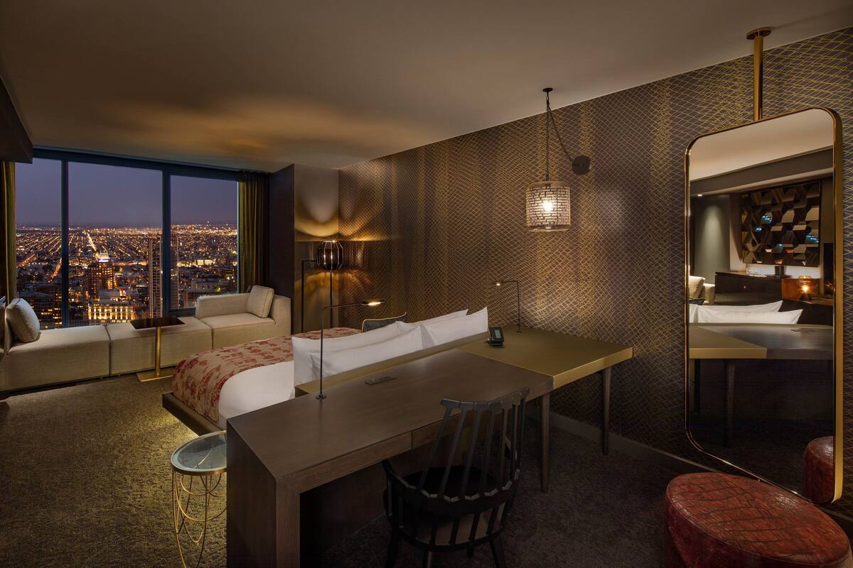 A modern hotel room with a city view, a large bed, a desk, and a seating area.
