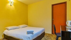 Ein Einzelbett mit weißer Matratze und Kissen, ein kleiner Tisch mit Stuhl, eine Tür und eine Wandleuchte.
