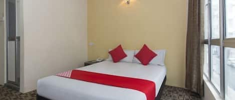 Une chambre d’hôtel comprenant un lit, une table de chevet, un appareil de climatisation, une fenêtre avec des rideaux et une lampe fixée au mur.