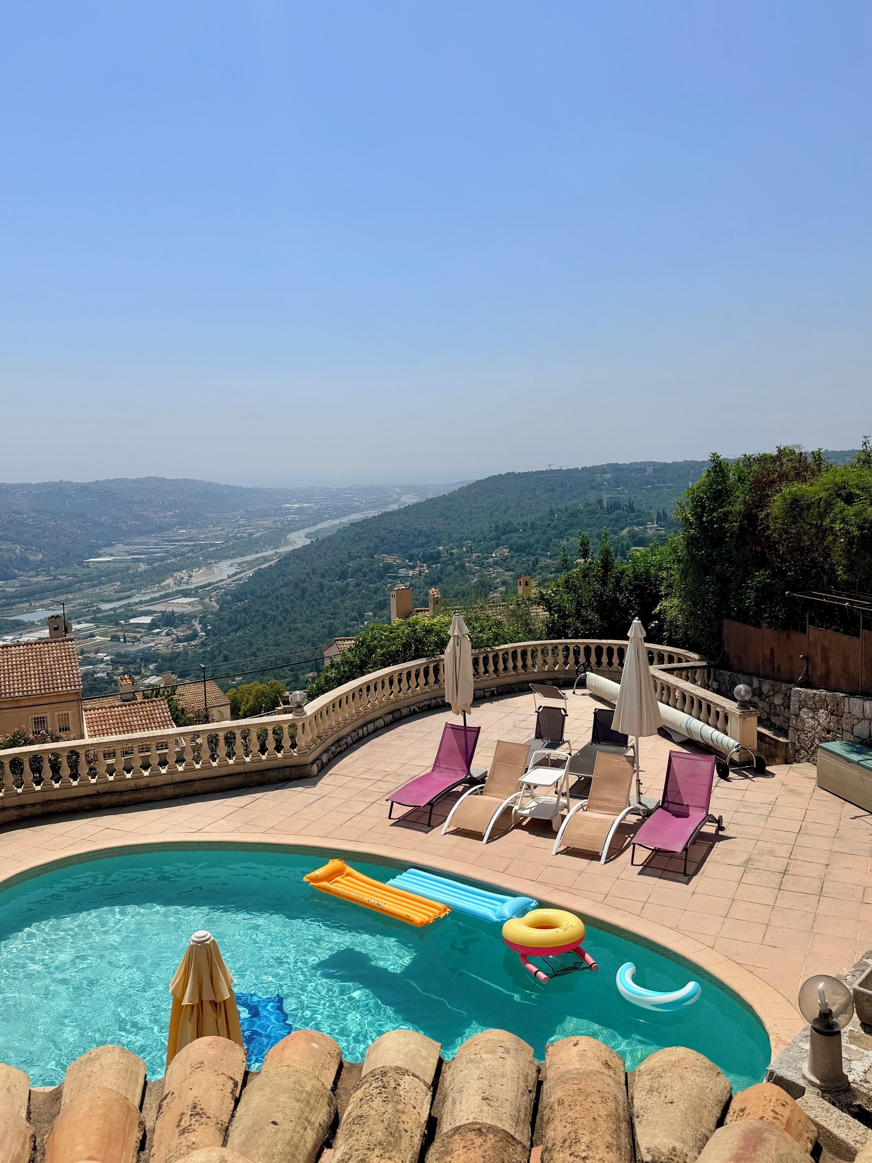 A pool area with lounge chairs and umbrellas, surrounded by a stone wall and overlooking a valley.