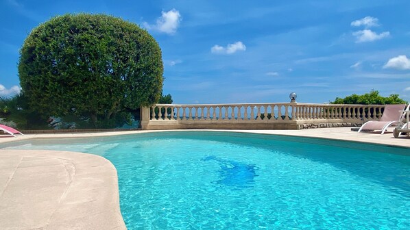 Piscina azul y cristalina con barandilla, rodeada de sillones de descanso y un árbol bien cuidado.