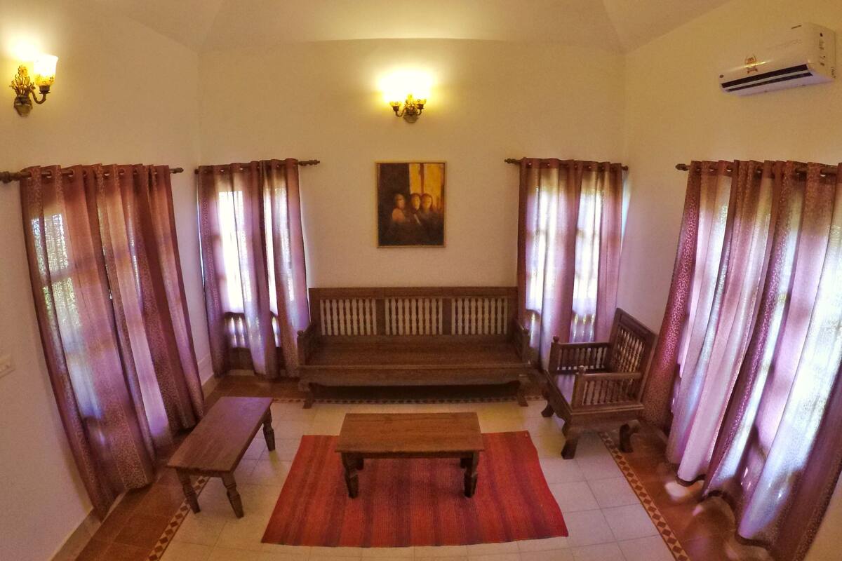 A living room with a wooden bench, a sofa, and a coffee table. There are curtains on the windows and an air conditioning unit on the wall.