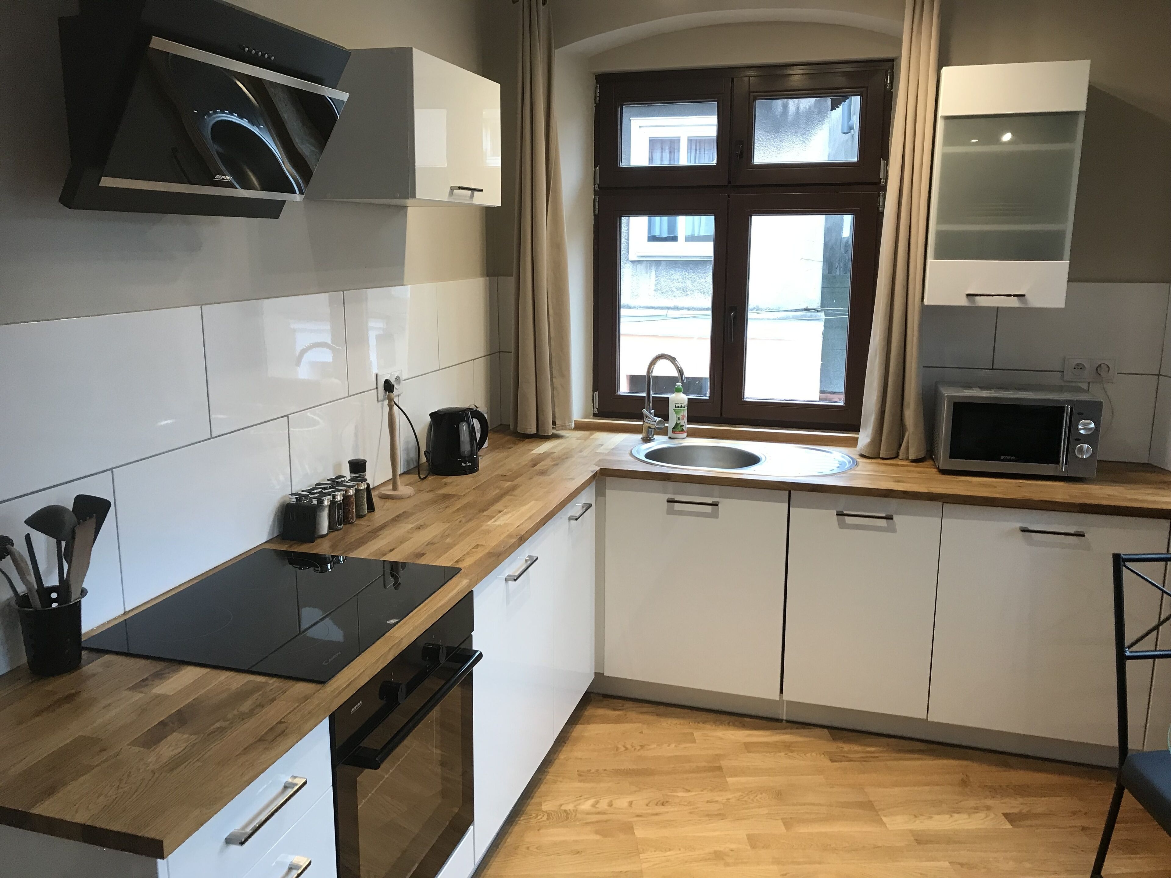 A modern kitchen with white cabinets, a wooden countertop, a built-in oven, a microwave, and a window with curtains.