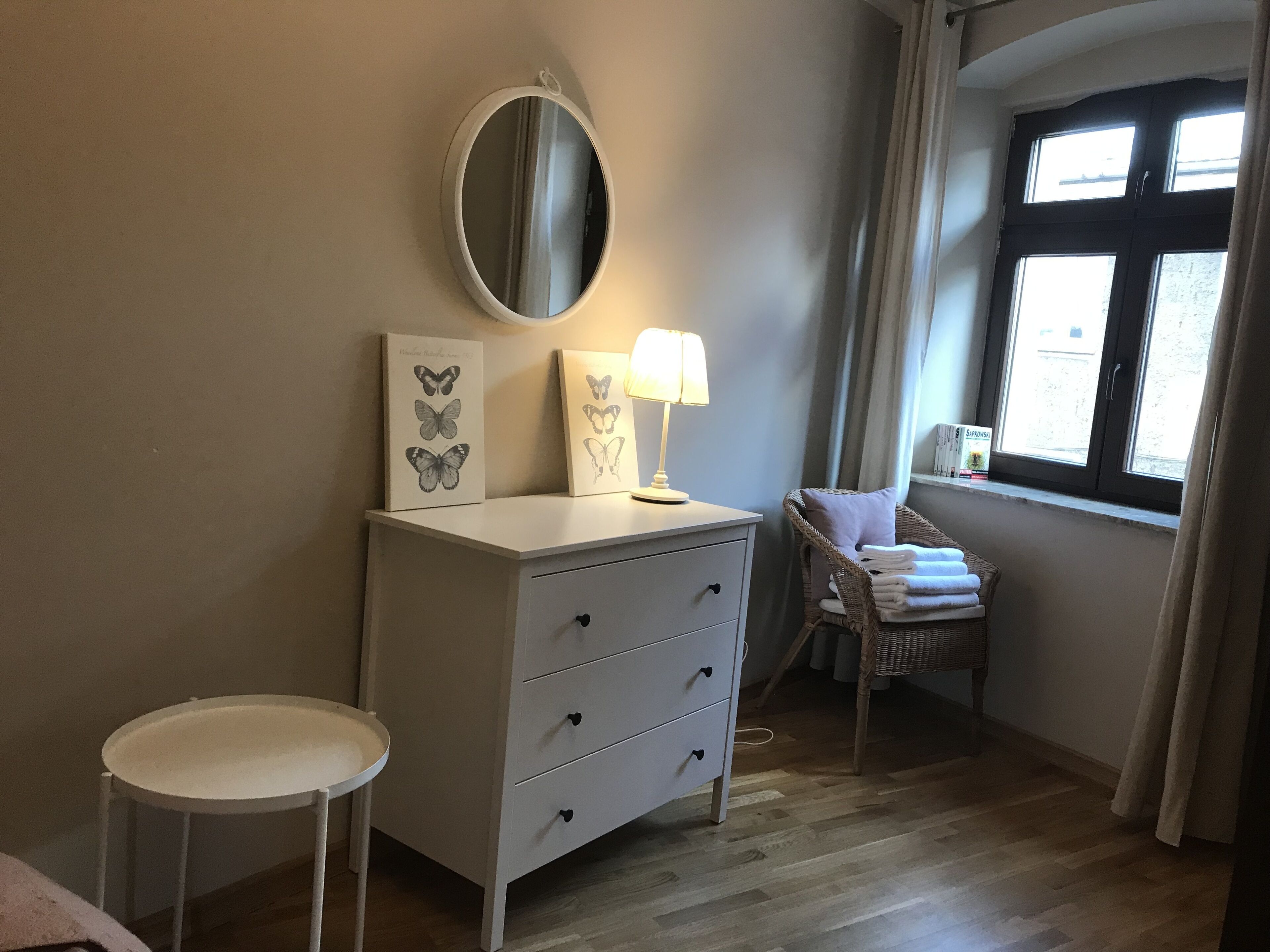 A bedroom with a dresser, a mirror, a bedside table, a chair, and a window with curtains.
