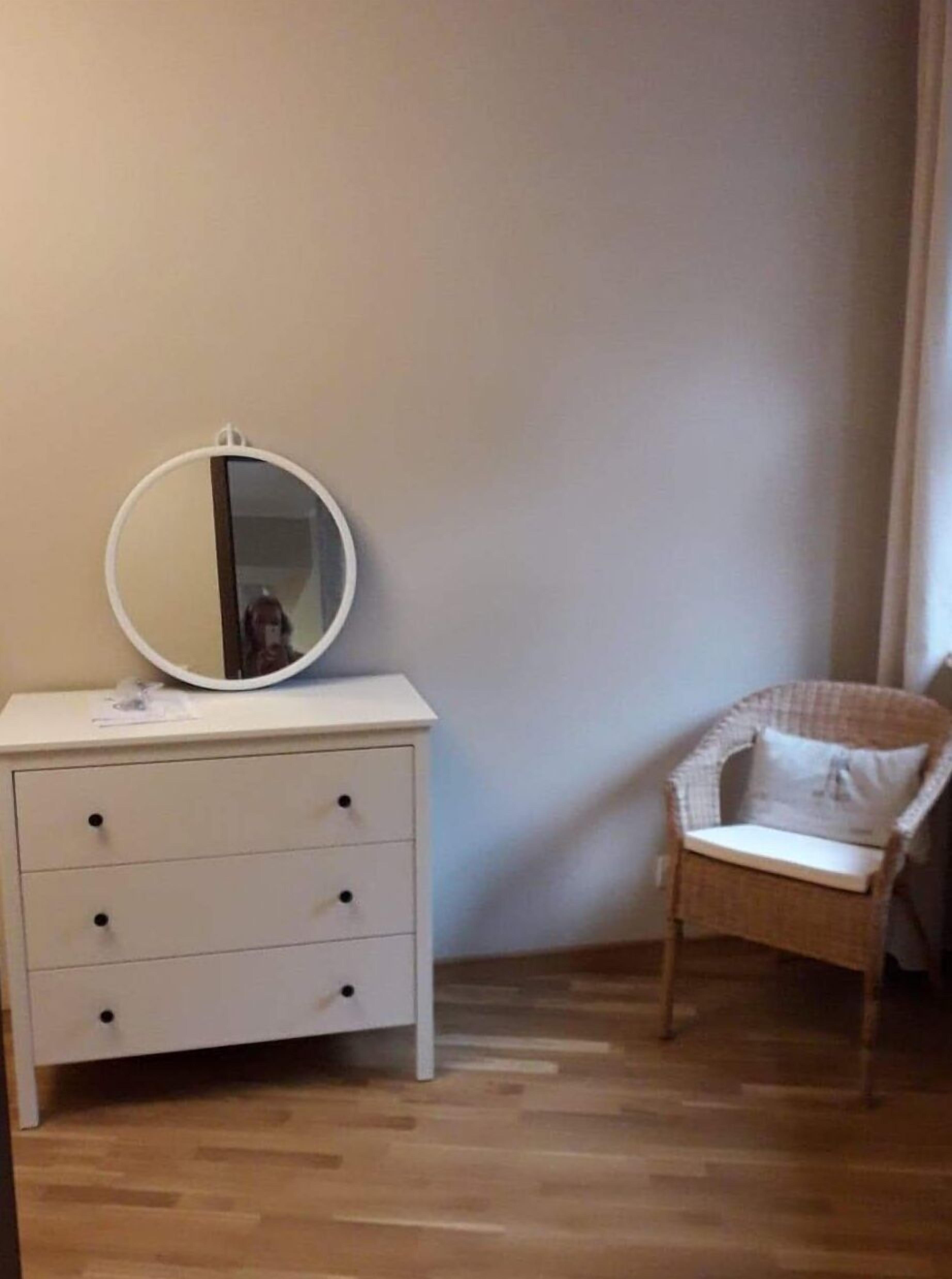 Ett rum med en vit byrå, en rund spegel och en fåtölj av rotting.