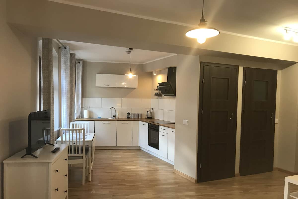 Una cocina compacta con armarios blancos, encimera de madera y horno empotrado. Cuenta con un comedor con mesa y sillas blancas, y un televisor sobre un mueble.