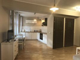 Una cocina compacta con armarios blancos, encimera de madera y horno empotrado. Cuenta con un comedor con mesa y sillas blancas, y un televisor sobre un mueble.