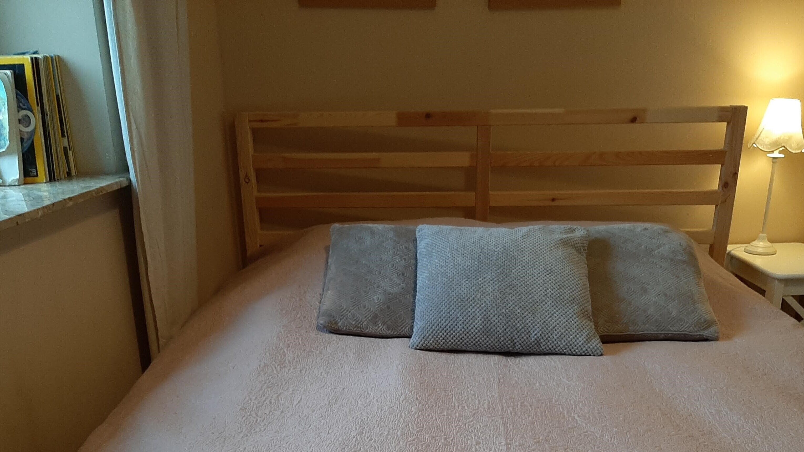 Een bed met een houten hoofdbord, twee kussens, een bedlampje en een plank met boeken en een fotolijst.