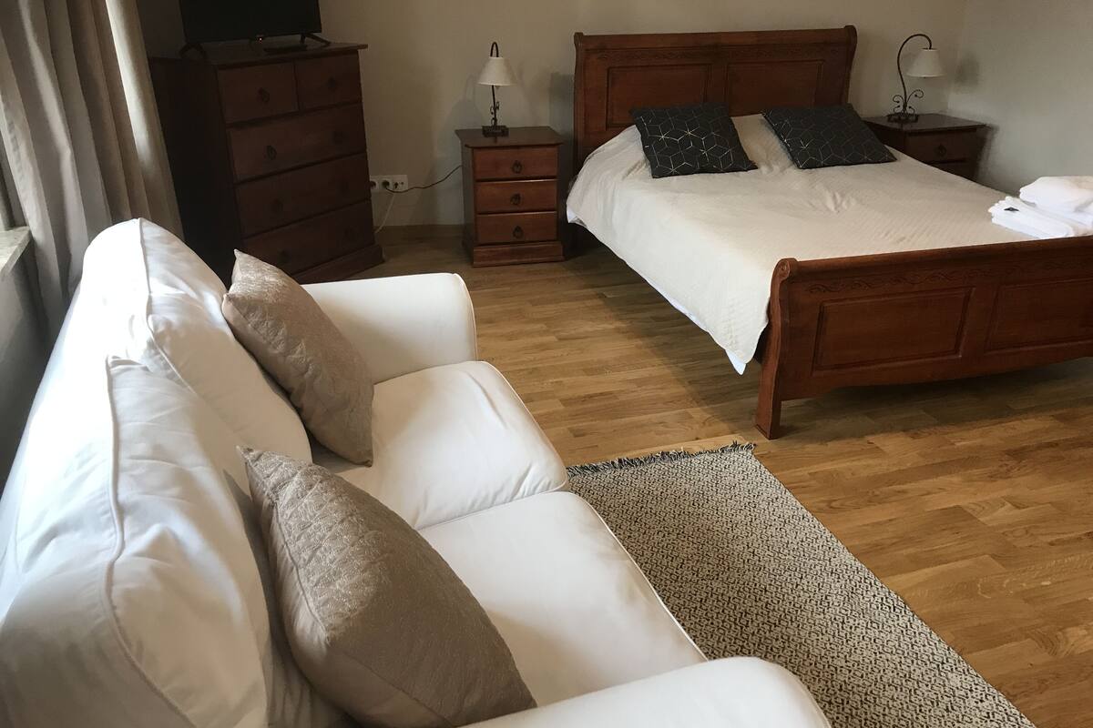 Un dormitorio con cama de madera, sofá blanco con cojines color beige, cómoda de madera y mesita de noche con lámpara.