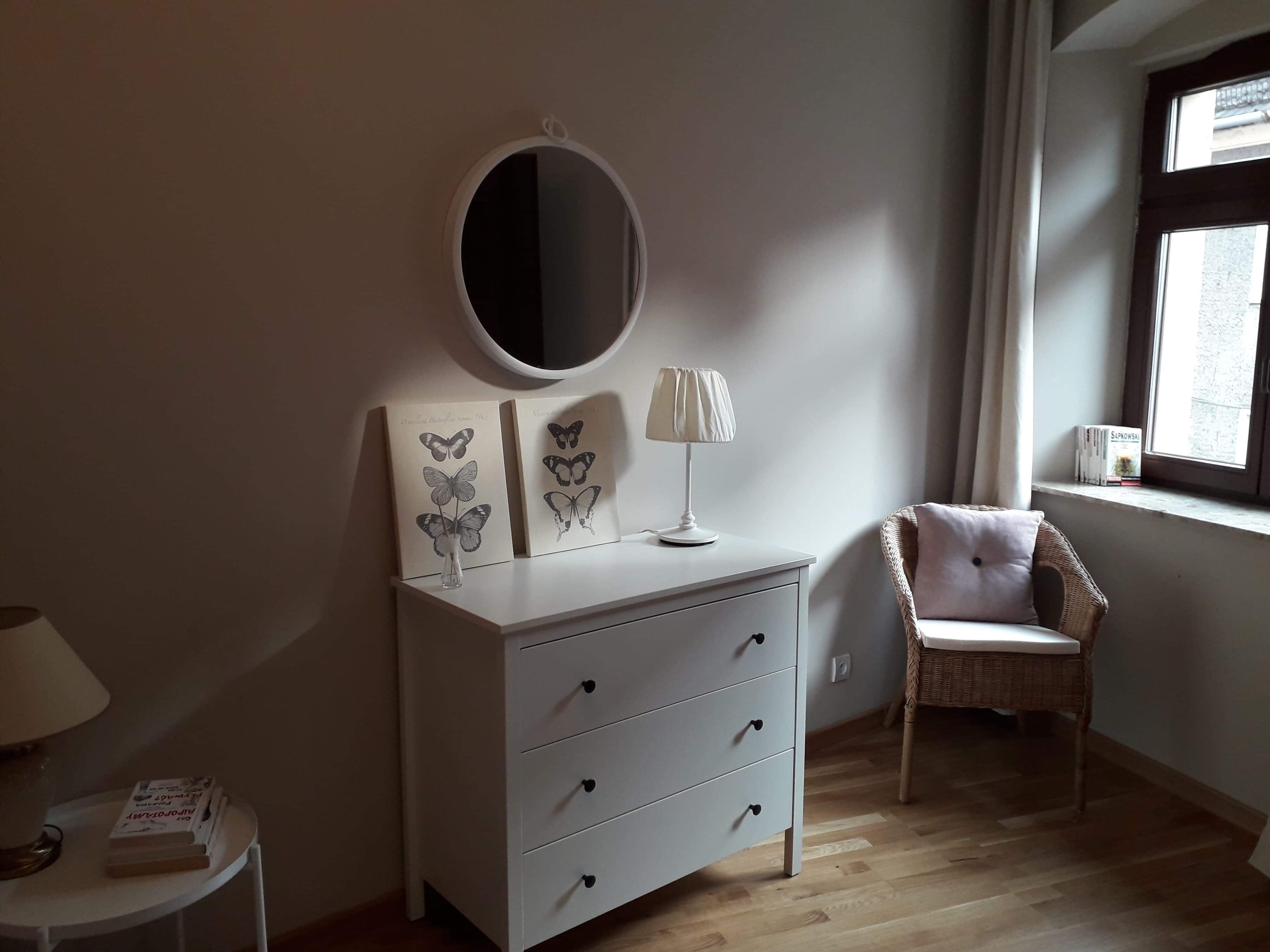 A bedroom with a white dresser, a round mirror, a lamp, and a wicker chair.