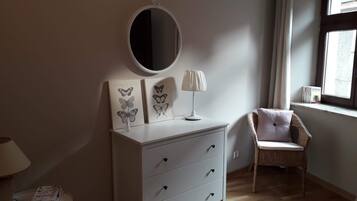 A bedroom with a white dresser, a round mirror, a lamp, and a wicker chair.