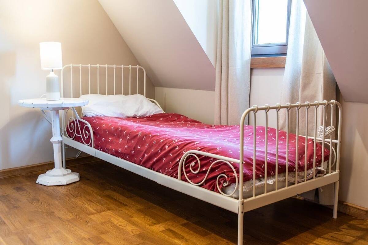 Una cama individual con estructura metálica blanca, colcha roja y mesita de noche con lámpara.