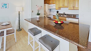 Une cuisine moderne avec un plan de travail en granit, des armoires en bois et un panier de fruits. On y trouve deux tabourets de bar blancs et une table à manger avec des chaises.