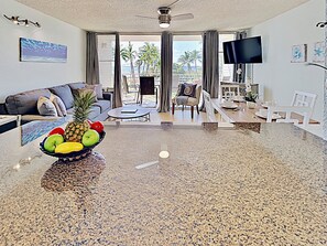 A modern living room with a granite countertop, a bowl of fruit, a sofa, a coffee table, and a dining area with a TV.