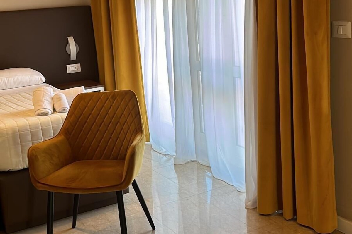 A hotel room with a bed, a chair, and a large window with sheer curtains.