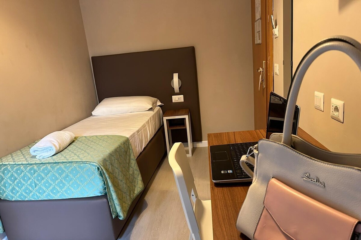 A hotel room with a bed, a desk with a laptop, a chair, a handbag, and a mirror.
