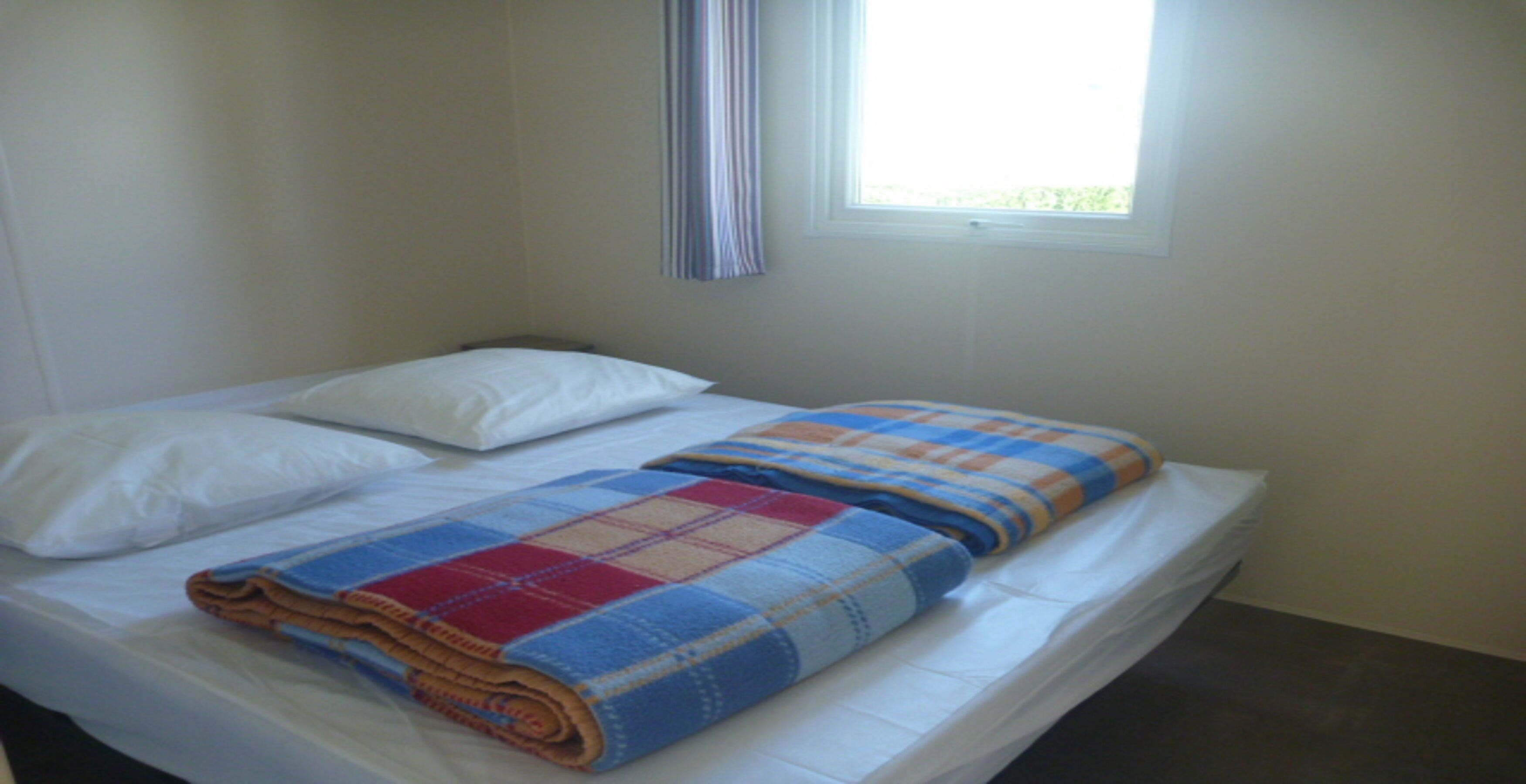 A single bed with white linens, a plaid blanket, and a window with vertical blinds.