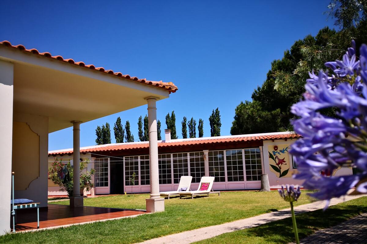 A building with a covered patio area, a swimming pool, and a garden with a pathway.