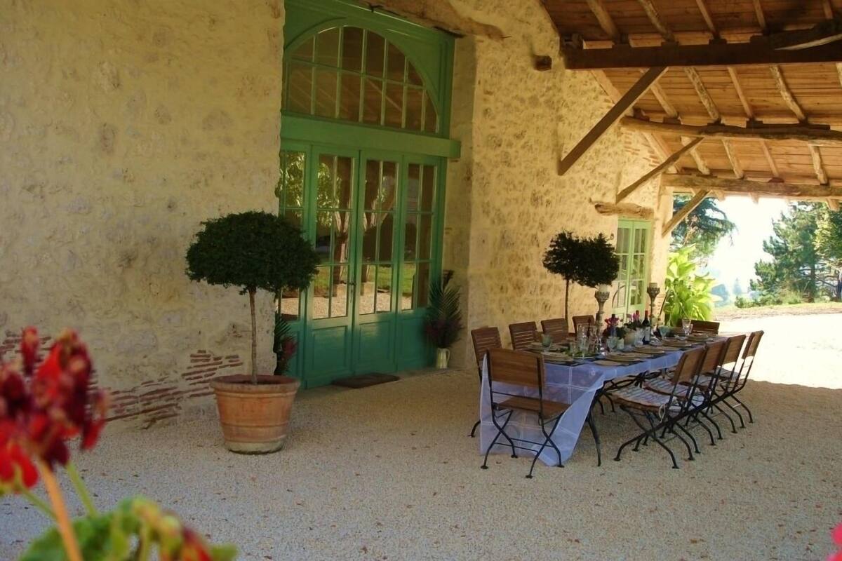 Un área de comedor al aire libre con una mesa dispuesta para seis personas, rodeada de sillas, bajo una pérgola de madera.
