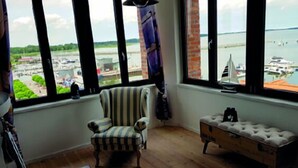 Een kamer met een groot raam dat uitkijkt op een jachthaven, een gestreept fauteuil, een bankje en een klein model zeilboot.