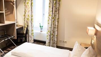 A hotel room with a bed, a desk, a chair, a radiator, and a window with curtains.