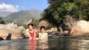 Two individuals in a river with large rocks and mountains in the background. - Serene Suites (Dharamshala)