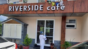 A Riverside Café with a wooden facade, a glass door entrance, and potted plants in front. - Serene Suites (Dharamshala)