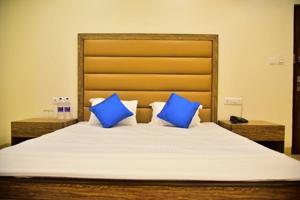 A hotel room with a large bed, two blue pillows, a wooden headboard, and a nightstand with a phone and water bottles.