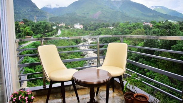 A balcony with two chairs and a table, overlooking a mountainous landscape with a river and buildings. - Serene Suites (Dharamshala)
