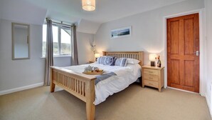 A bedroom with a wooden bed, bedside tables, a nightstand, a wooden door, a window with curtains, and a ceiling light fixture. - 5 Bed in Croyde (oc-fircon) (Croyde)