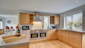 A modern kitchen with wooden cabinets, a central island, a microwave, a toaster, and a window with a view of greenery. - 5 Bed in Croyde (oc-fircon) (Croyde)