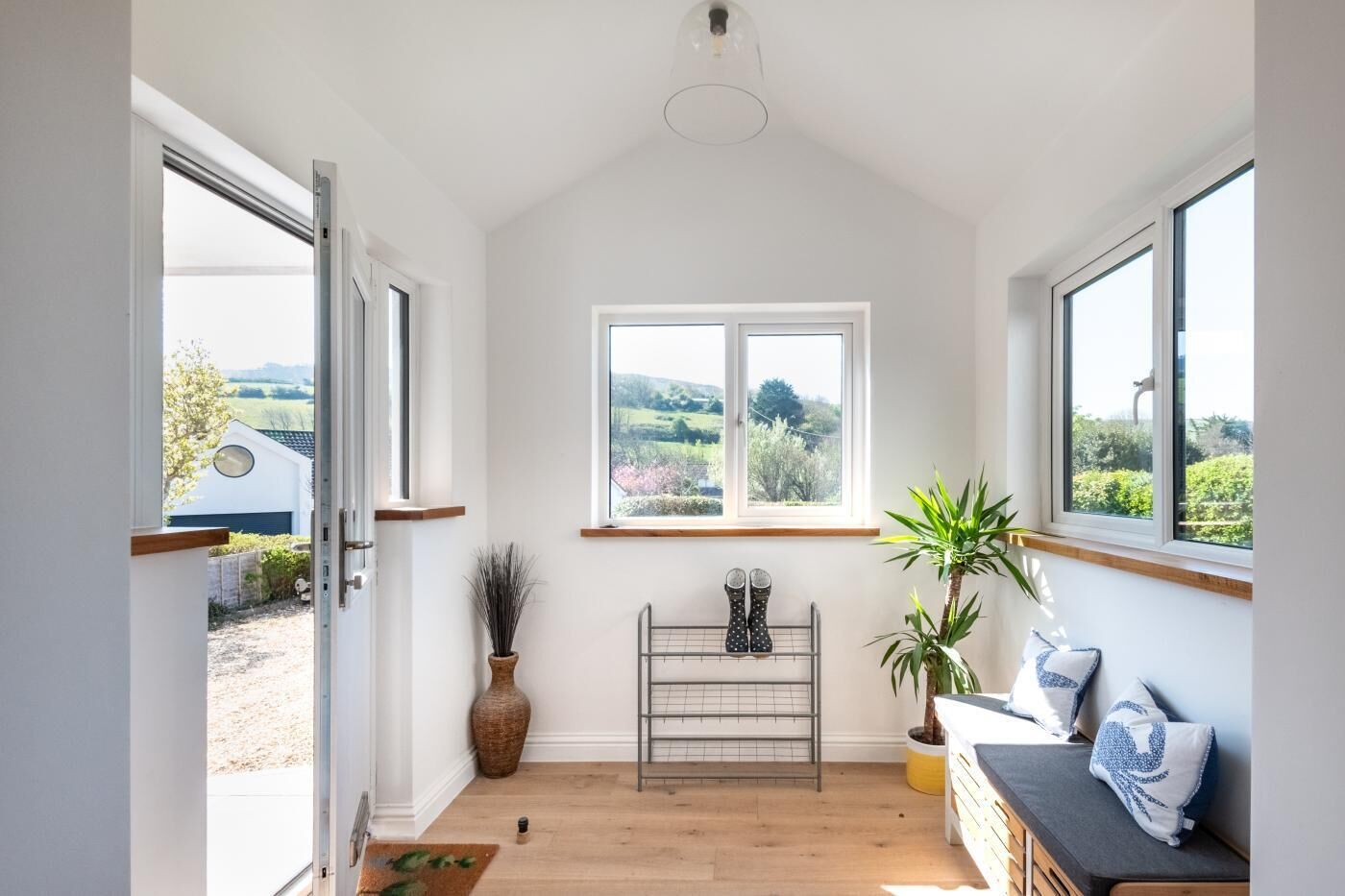A room with a wooden floor, a window with a view of greenery, a plant, and a bench.