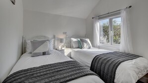 A bedroom with two beds, white bedding, a window with curtains, and a small bedside table with a lamp. - 3 Bed in Croyde (oc-moonra) (Croyde)