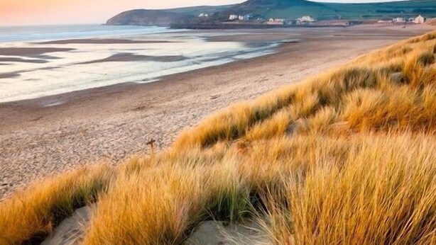 A coastal landscape with a sandy beach, grassy dunes, and a distant hill with buildings. - 3 Bed in Croyde (oc-moonra) (Croyde)