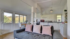 A cozy living room with a grey sofa, a dining area with a table and chairs, and a kitchen with white cabinets and appliances. - 3 Bed in Croyde (oc-moonra) (Croyde)