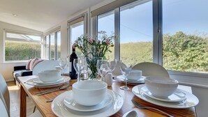 A dining area with a table set for four, large windows, and a view of greenery outside. - 3 Bed in Croyde (oc-moonra) (Croyde)