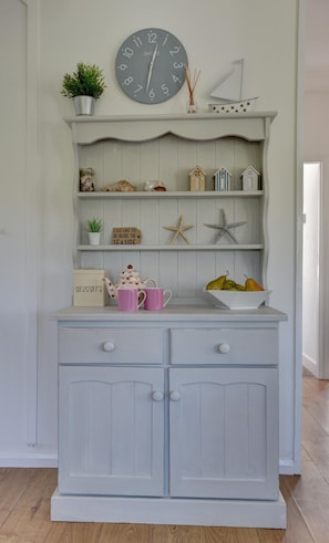 A light blue wooden cabinet with two drawers and a shelf above. On the shelf, there is a clock, a small potted plant, a jar, a sign, a starfish, and a bowl of fruit. - 3 Bed in Croyde (oc-moonra) (Croyde)