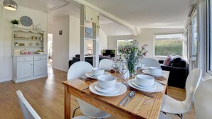 A dining area with a wooden table set for four, white chairs, and a view of the outdoors through large windows. - 3 Bed in Croyde (oc-moonra) (Croyde)