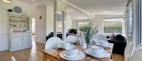 Un coin repas avec une table en bois dressée pour quatre personnes, des chaises blanches et une vue sur l’extérieur grâce à de grandes fenêtres.