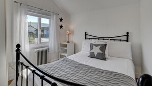 A bedroom with a bed, a nightstand, a lamp, a window with curtains, and a radiator. - 3 Bed in Croyde (oc-moonra) (Croyde)