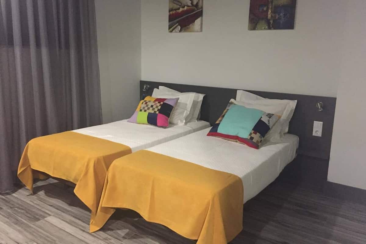 Ein Hotelzimmer mit zwei Betten, jeweils mit einer gelben Tagesdecke bezogen. Der Boden ist aus Holz und das Fenster hat einen grauen Vorhang. Über den Betten hängen zwei Bilder an der Wand.