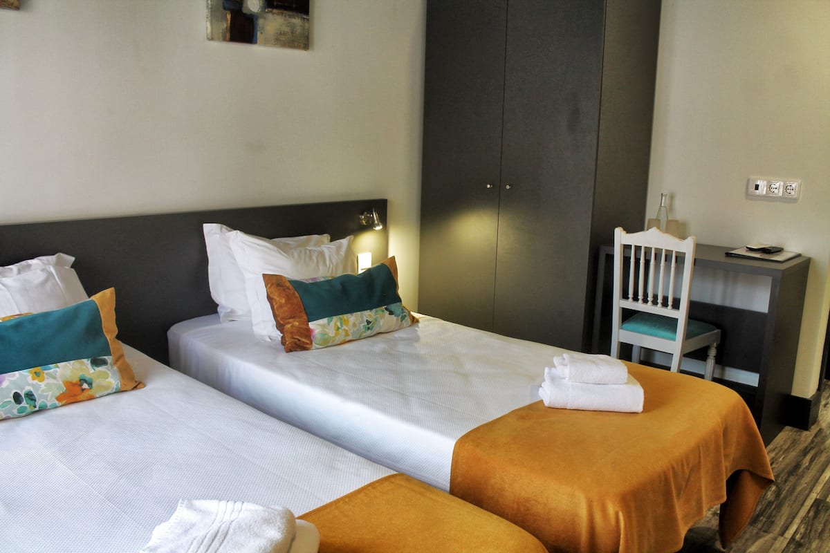Ein Hotelzimmer mit zwei Betten, jeweils mit weißer und orangefarbener Bettdecke, einem Nachttisch mit Lampe und einem Schreibtisch mit Stuhl.