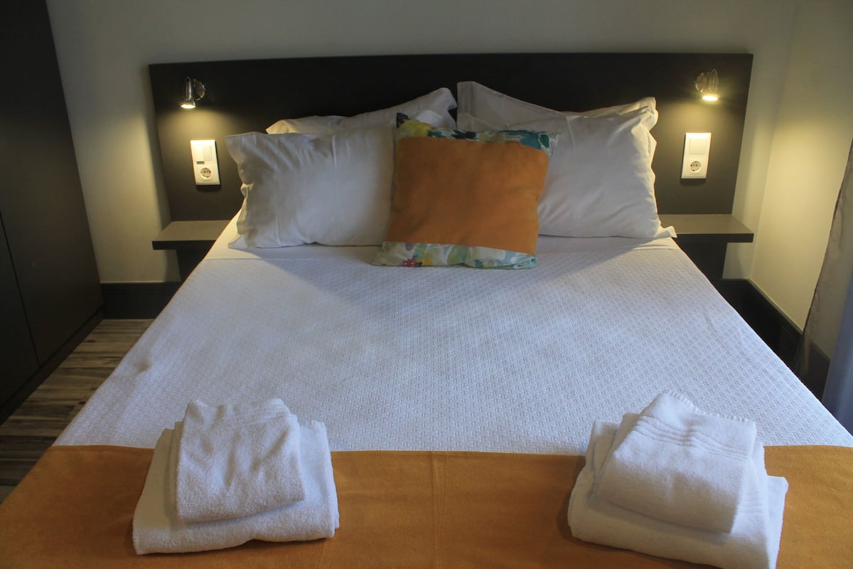 Ein ordentlich bezogenes Bett mit weißer Bettwäsche und einem einzelnen orangefarbenen Kissen, zwei gefaltete Handtücher auf dem Bett und Wandleuchten an jeder Seite.