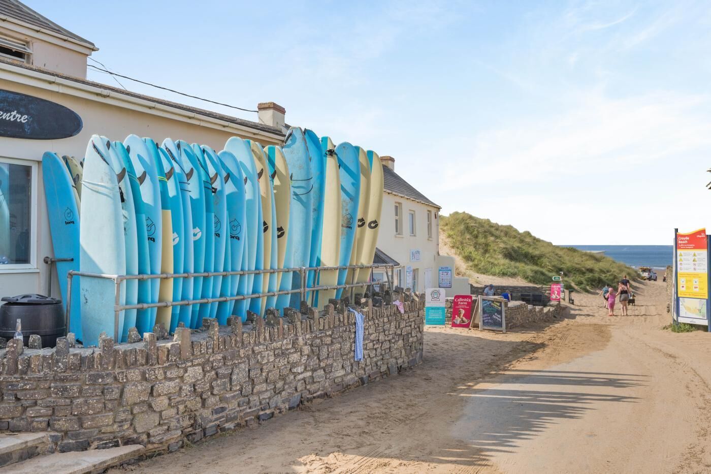 A coastal building with surfboards leaning against the wall, a sandy beach, and a clear sky.