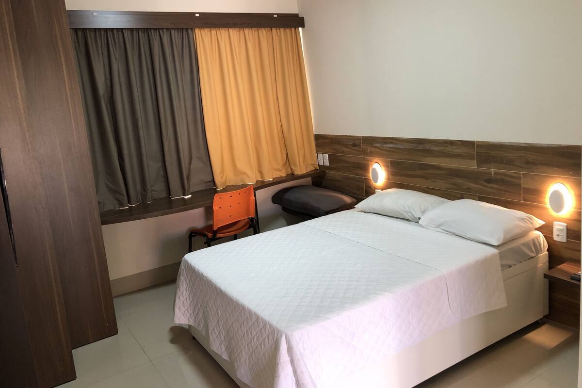 Una habitación de hotel con cama, una silla, una ventana con cortinas y una mesita de noche con lámpara.