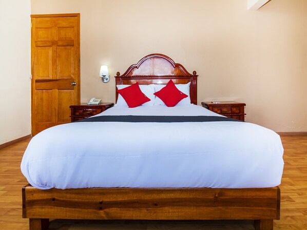 Una cama bien hecha con cabecera de madera, ropa de cama blanca y dos almohadas rojas. Se observa un mesita de noche de madera con una lámpara y un teléfono encima. Se ve una puerta de madera al fondo.