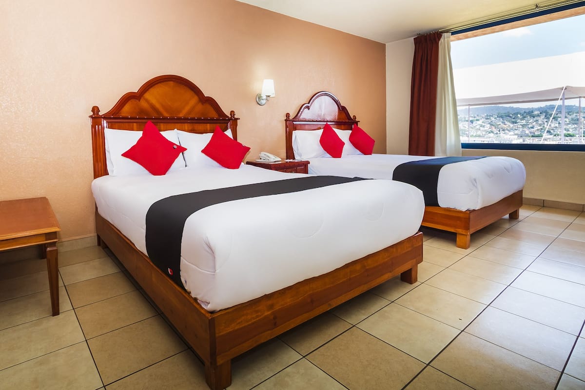 Une chambre d’hôtel avec deux lits en bois, du linge de lit blanc, des oreillers rouges, une table de chevet, une commode, une lampe et une fenêtre donnant sur un beau paysage.